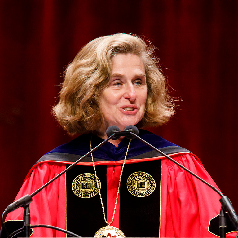 IU President Pamela Whitten in her formal regalia of doctoral gown and the university Jewel, speaking  at a lectern.